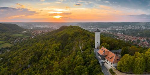 Blick aus der Vogelperspektive auf einen historischen Bergfried, der sich auf einem Berg befindet. Im dahinter gelegenen Tal erstreckt sich die Stadt Jena im Abendlicht.
